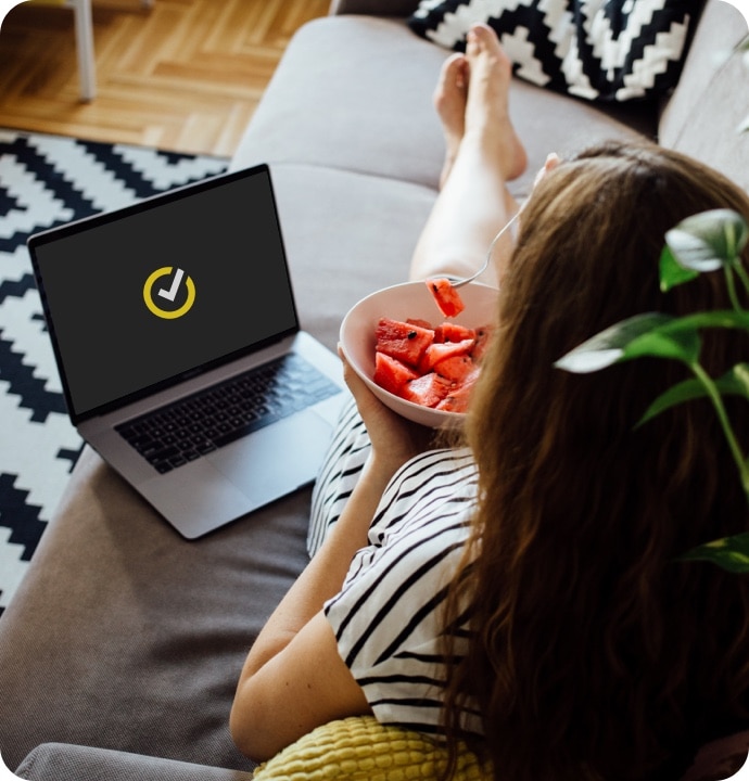 Mulher a comer melancia no sofá com um portátil a iniciar o Norton 360 AntiVirus Plus